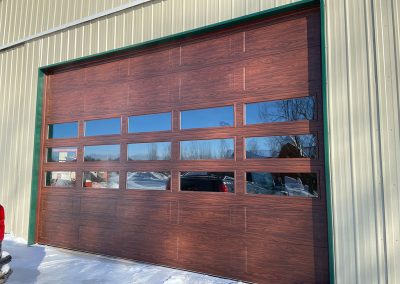 A commercial building with a brown garage door. In the reflection of the garage door mirror, there is a snow-covered environment and a car.